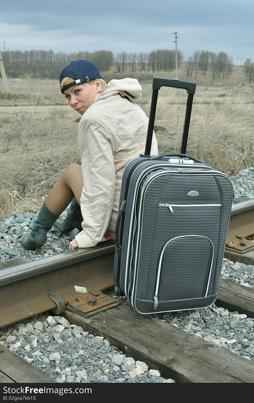 She is sitting with a bag on the rails, in a light jacket and a cap for baseball, turned her head. She is sitting with a bag on the rails, in a light jacket and a cap for baseball, turned her head