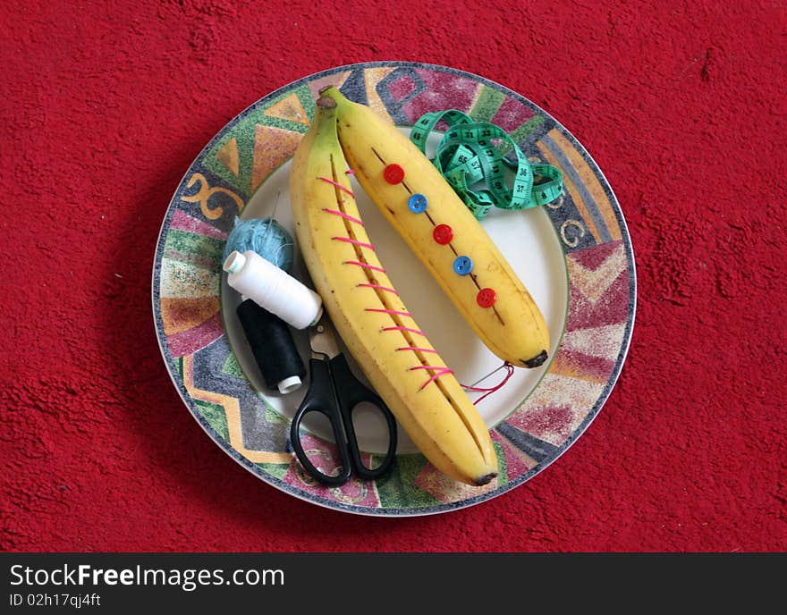 Two bananas on a plate, thread, scissors, a centimeter tape, buttons, a needle