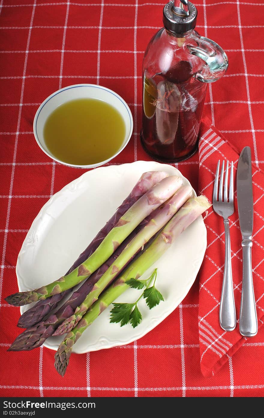 Bunch of fresh organic and home-grown asparagus on a white dish resting on a red table cloth  with a bottle of vinegar in the background next to a small bowl of olive oil. Folded napkin and silverware on top. Bunch of fresh organic and home-grown asparagus on a white dish resting on a red table cloth  with a bottle of vinegar in the background next to a small bowl of olive oil. Folded napkin and silverware on top.