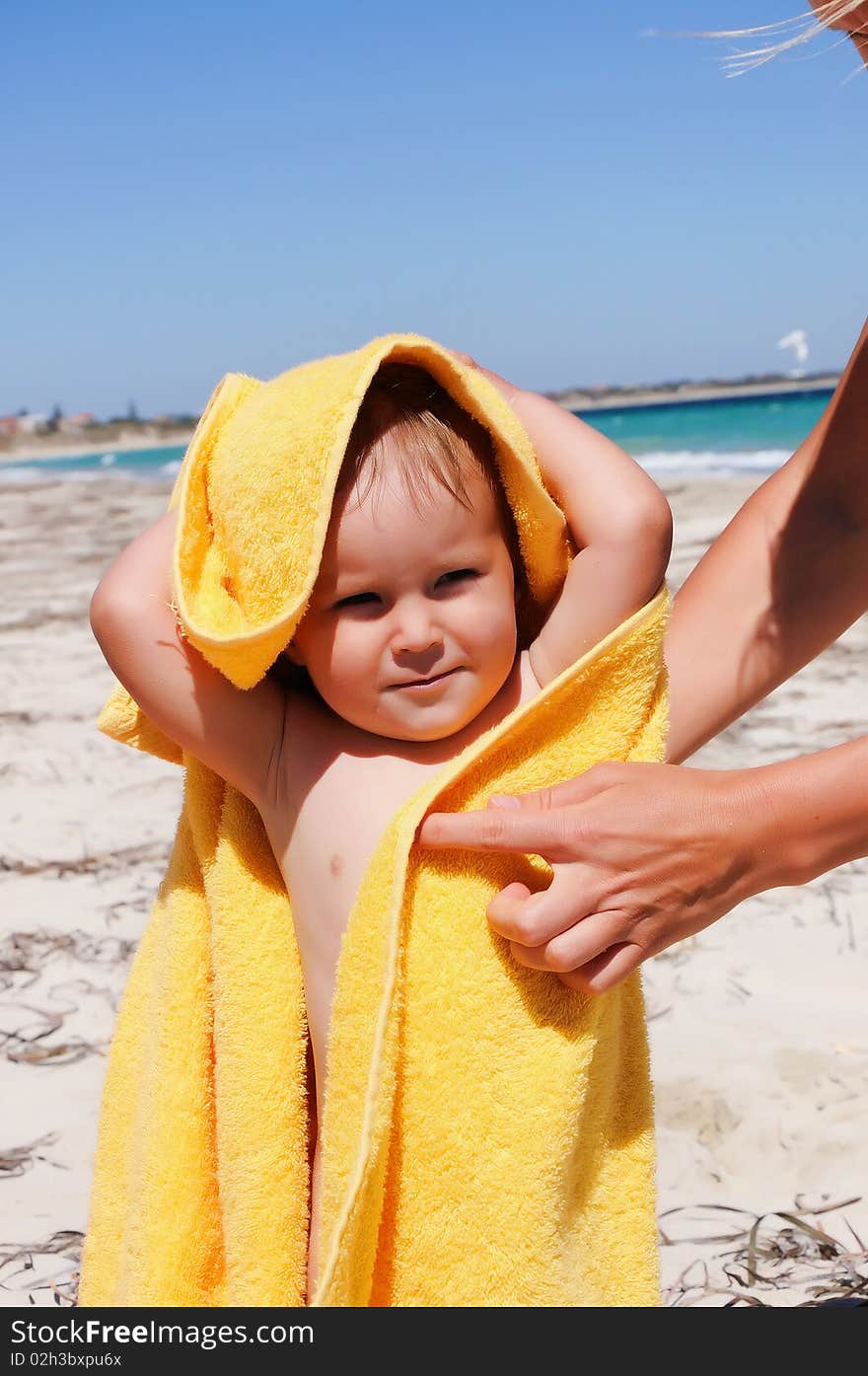 Smiling little girl in a yellow towel on the beach. Caring mother's hands to help her.