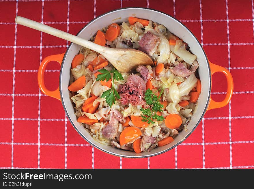 Large pot filled with ham, cabbage and carrots and parsley. The table is dressed with a checkered red cloth. Large pot filled with ham, cabbage and carrots and parsley. The table is dressed with a checkered red cloth.