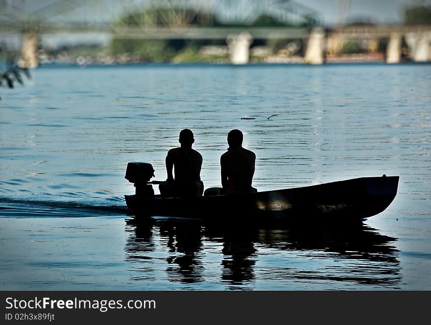 A two man in a boat, afternoon light silhouettes, bridge in out on focus background