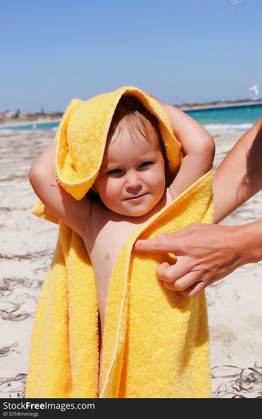 Smiling little girl in a yellow towel on the beach. Caring mother's hands to help her.