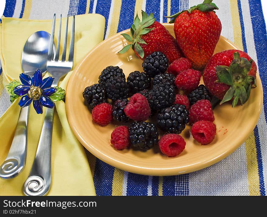 Assorted berries on a yellow plate on a striped textile with napkin, silverware, and napkin ring.