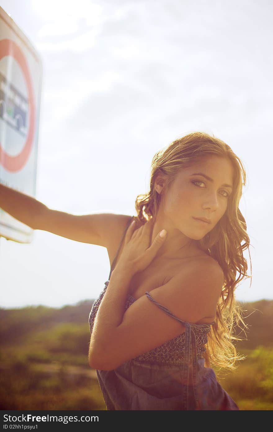 Beautiful fashion female model with blond hair looking at the camera and holding on a road sign. Photo taken on sunset. Beautiful fashion female model with blond hair looking at the camera and holding on a road sign. Photo taken on sunset