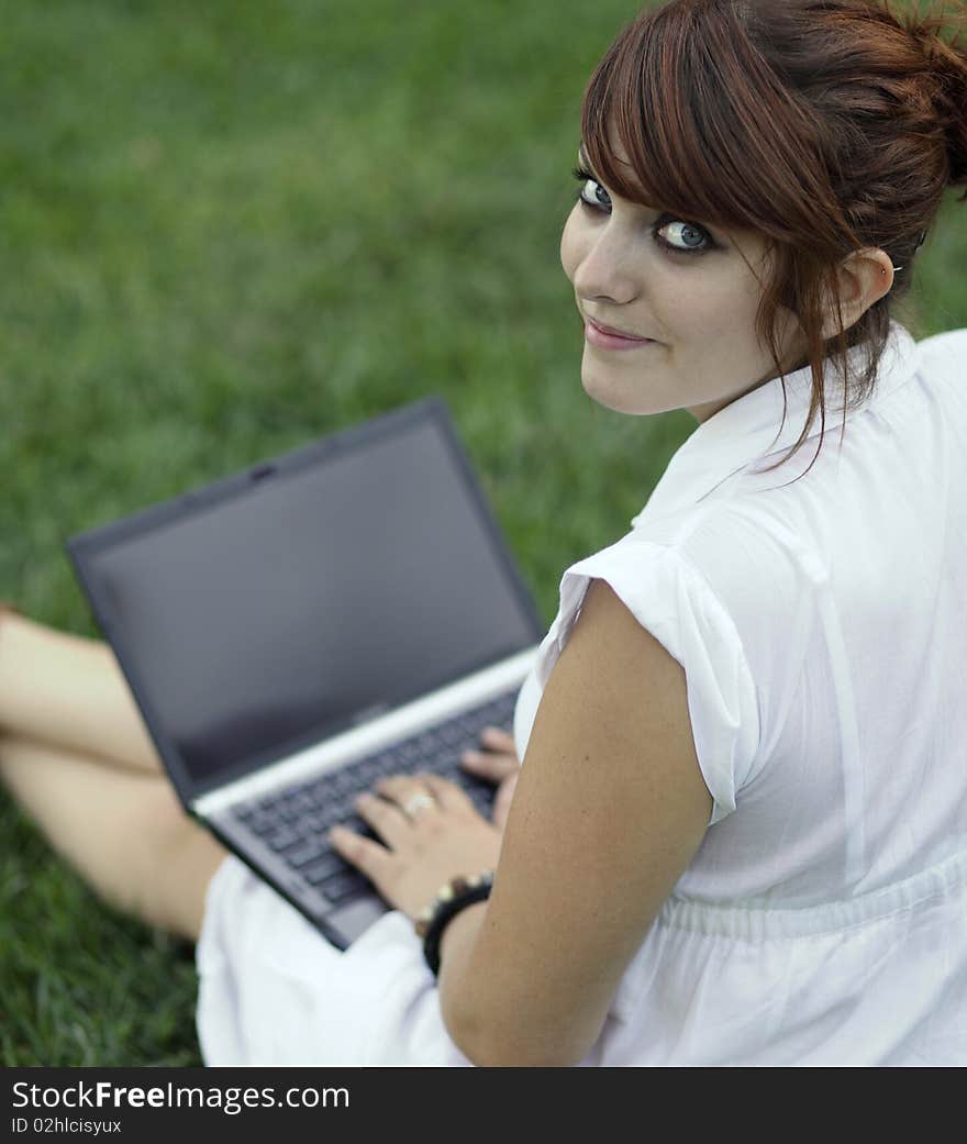 Pretty young woman working on a laptop computer outdoors, lying on green grass, pondering, looking away