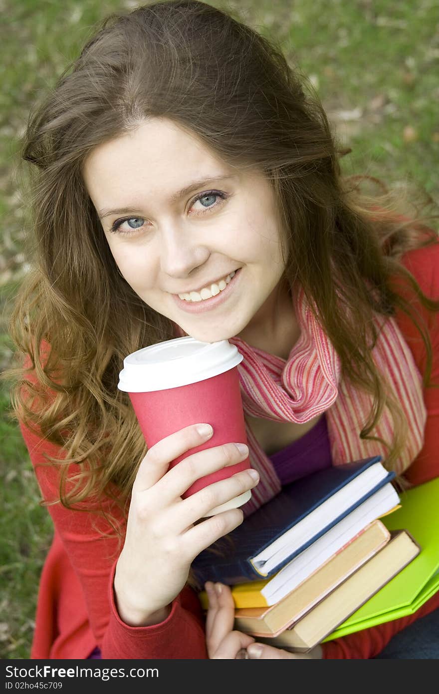 Young beautiful student at a dinner in the park sits on the green grass drinks coffee. Keep lots of books and folders. Young beautiful student at a dinner in the park sits on the green grass drinks coffee. Keep lots of books and folders