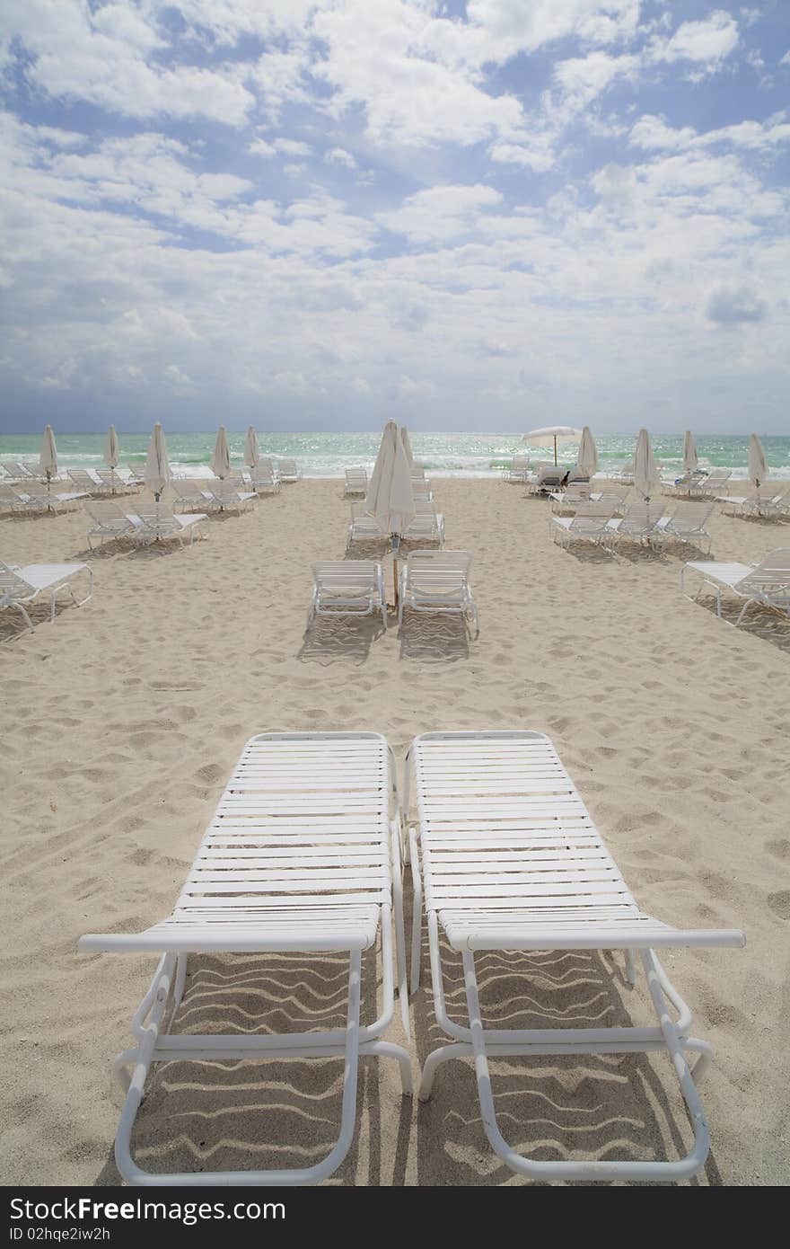 Rows of beach lounge chairs located in Miami's South Beach area in the morning sunlight with blue sky and clouds above and ocean in the background.