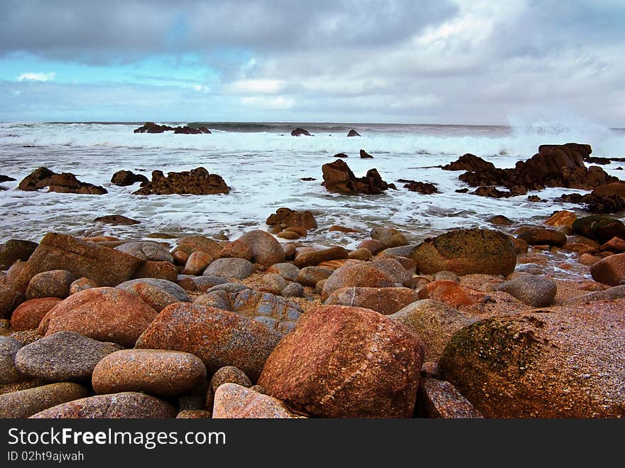 A wide shot showing colorful boulders with large waves crashing in the background. A wide shot showing colorful boulders with large waves crashing in the background