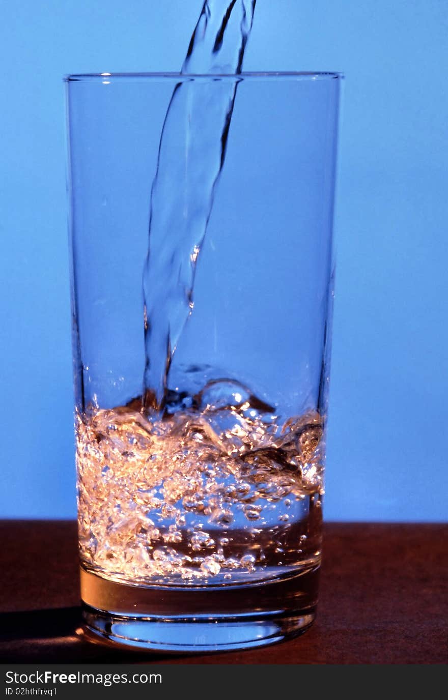 Water is being poured into glass against a blue background. Vertical shot. Water is being poured into glass against a blue background. Vertical shot.