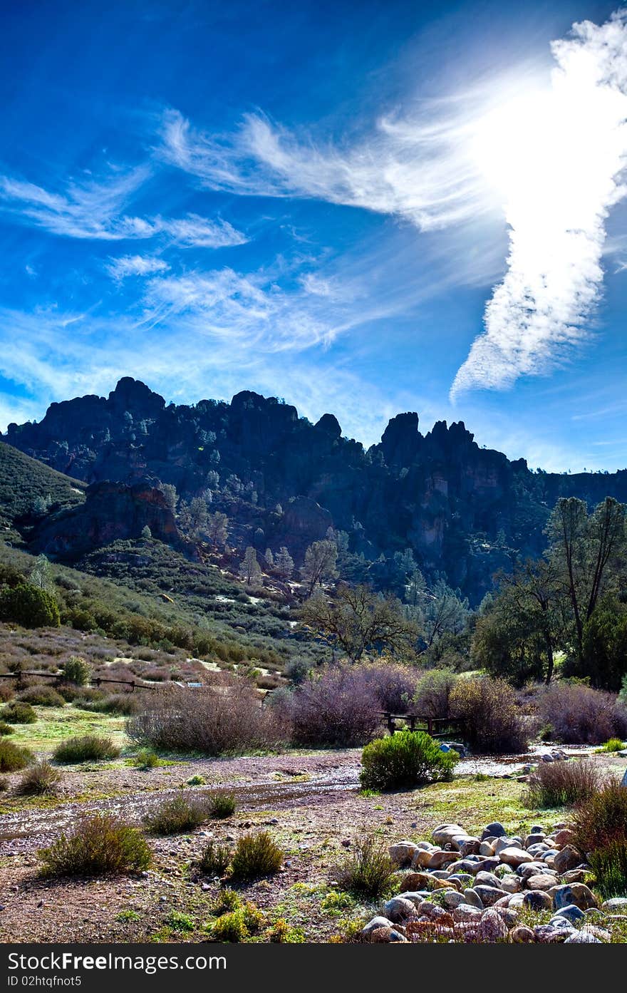 Mountain range in distance, grass and trees. Sky is filled with thine clouds and sun is out. Creek running across. Mountain range in distance, grass and trees. Sky is filled with thine clouds and sun is out. Creek running across