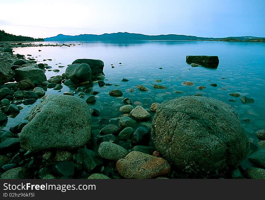 Boulders, cobble, rocks and stones on a beach are exposed at low tide in a calm inlet with distant shore and morning sky background. Boulders, cobble, rocks and stones on a beach are exposed at low tide in a calm inlet with distant shore and morning sky background.