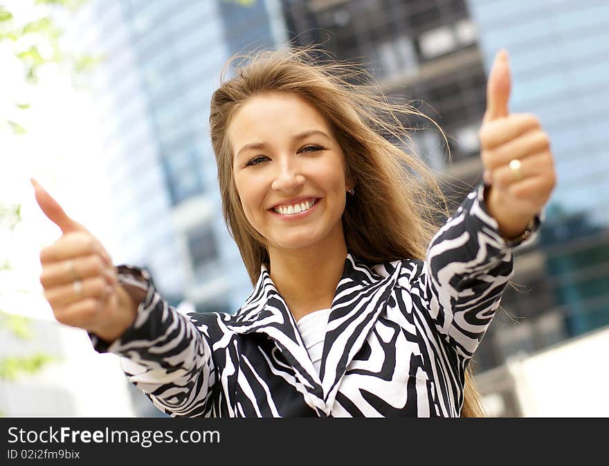 Portrait on a young business woman holding thumbs up as a sign of success. Image taken on a outdoors, on a modern background.