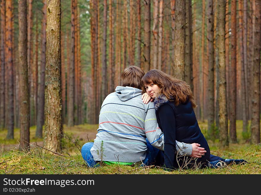 Young loving couple sitting back on a grass in the forest, woman is turning her face, they are hugging each other and dreaming. Young loving couple sitting back on a grass in the forest, woman is turning her face, they are hugging each other and dreaming