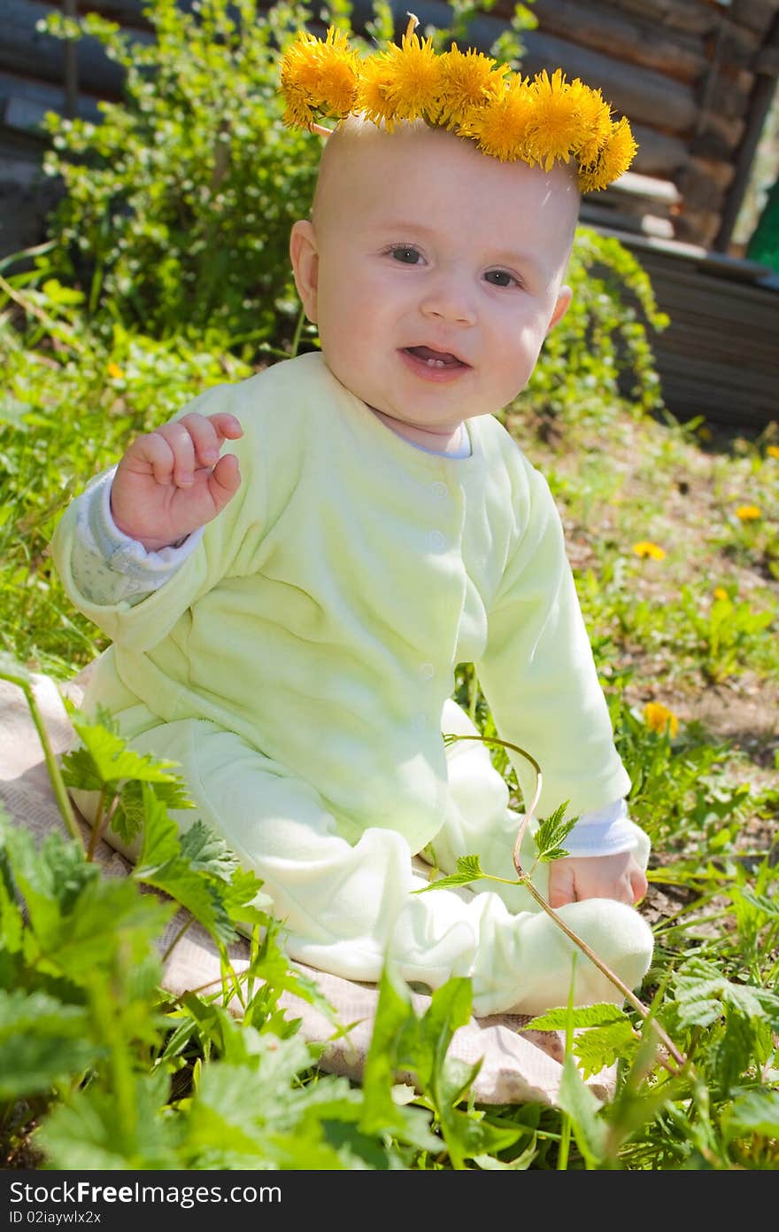 The baby of 7-8 months sits on a grass with a wreath from dandelions on a head and waves discontentedly hands