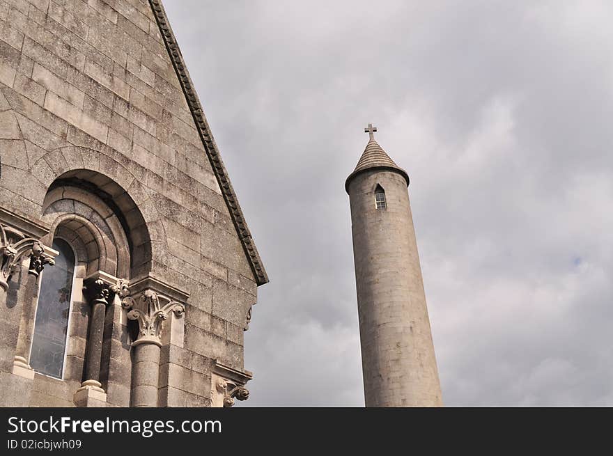 Image of a tall graveyard tower with window on top and section of small church with arch window in cemetery in north dublin ireland on cloudy day.