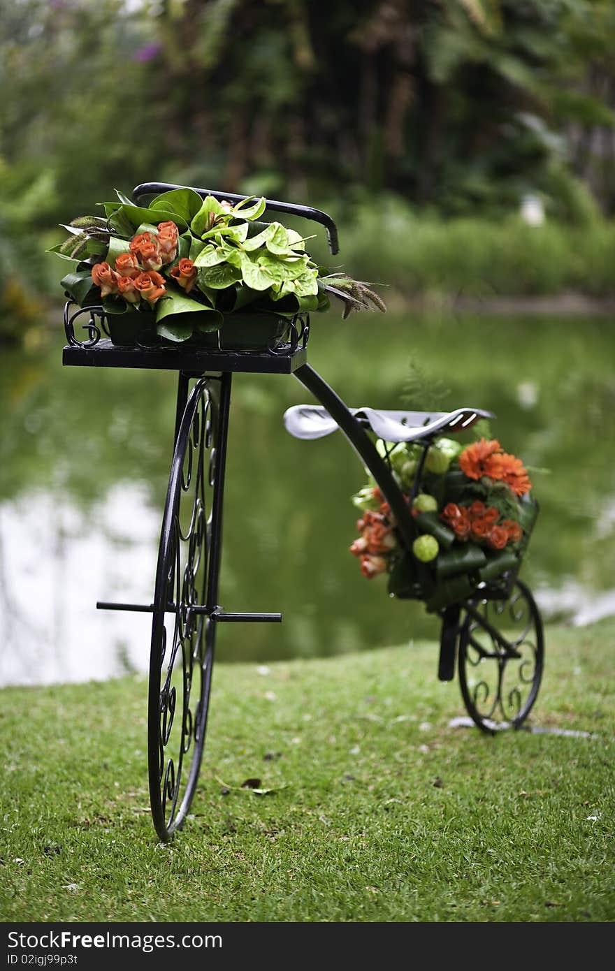 A wrought iron bicycle decorated with flowers on a green grass patch with a pond in the background. A wrought iron bicycle decorated with flowers on a green grass patch with a pond in the background