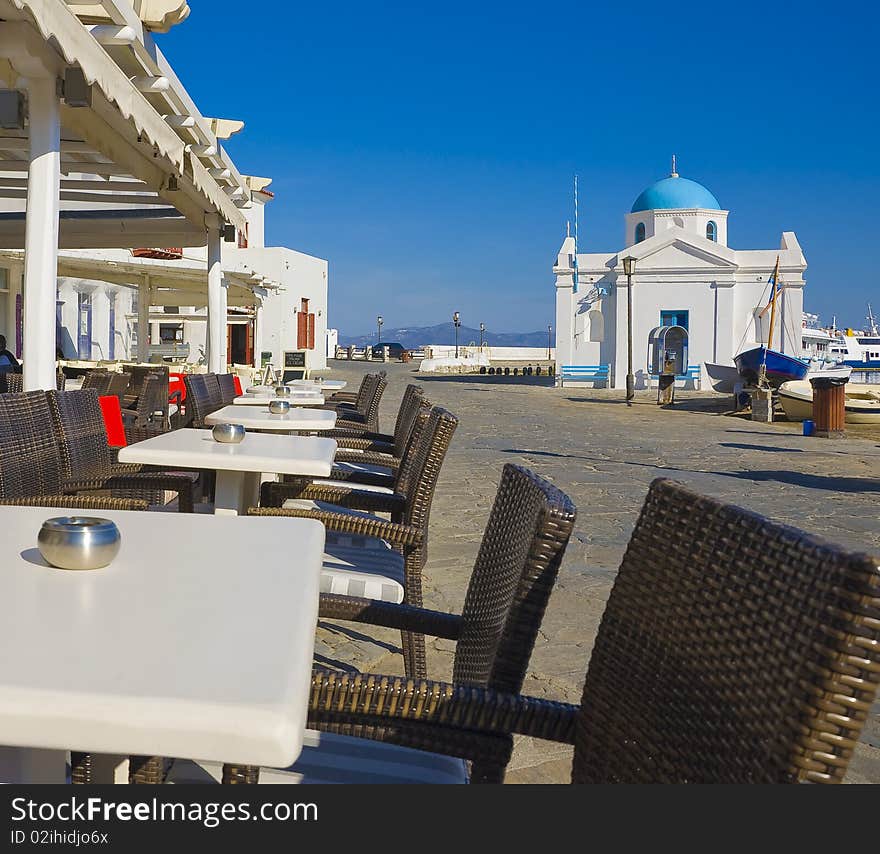 Tables and chairs for the restaurant on the seafront of Mykonos on the background of the church. Tables and chairs for the restaurant on the seafront of Mykonos on the background of the church