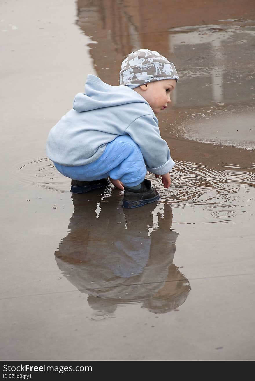 A little boy crouching down into a big puddle; with a reflection of a boy and a building. A little boy crouching down into a big puddle; with a reflection of a boy and a building.