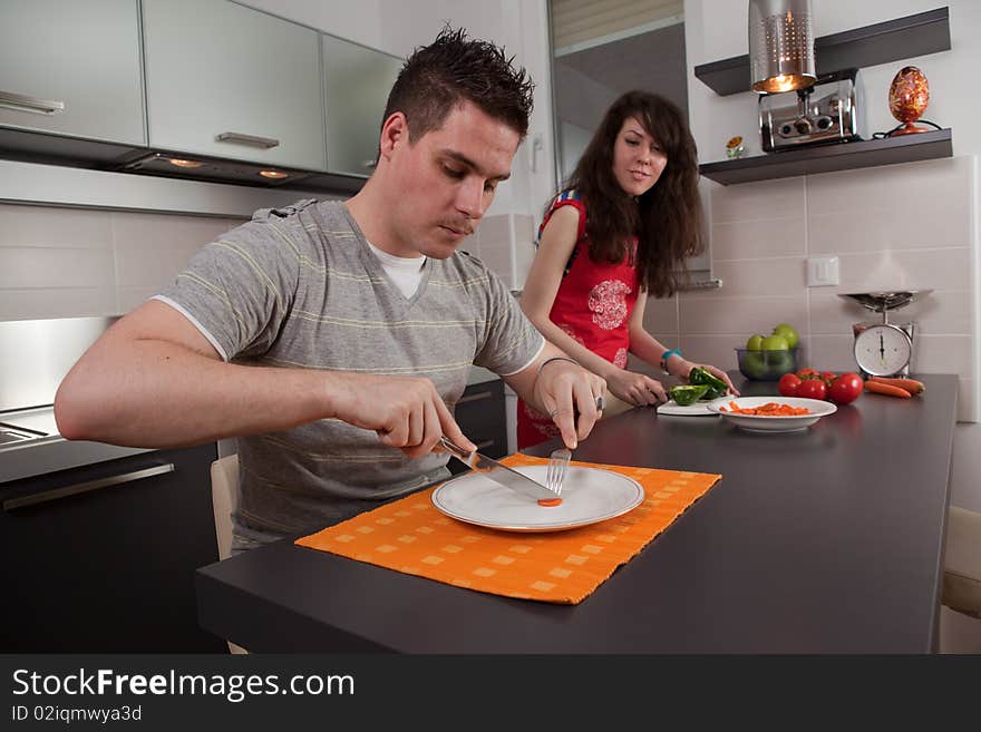 Hungry guy eating a slice of carrot while girl cooks.