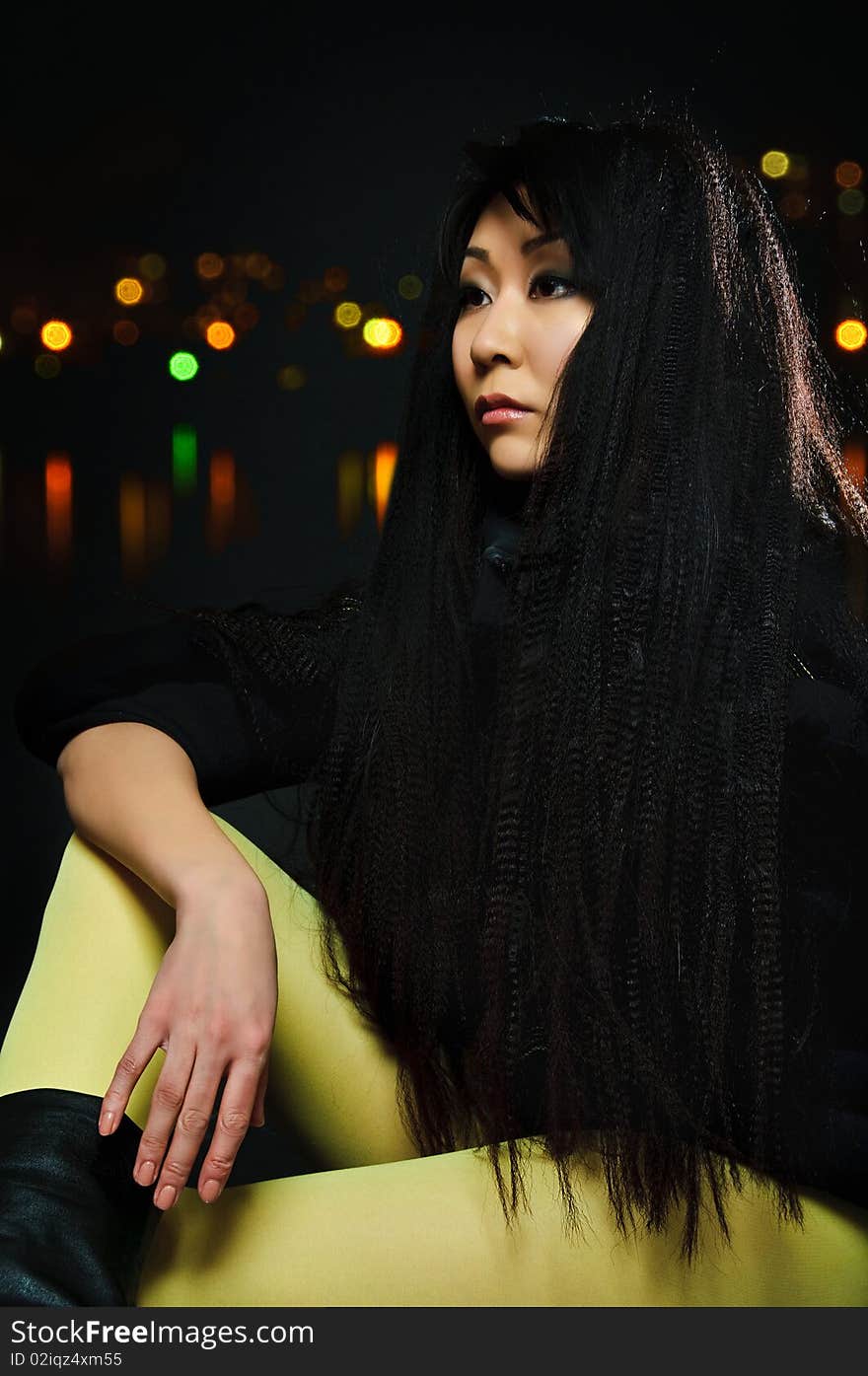 Sitting brunette asian model on black background with night light reflections