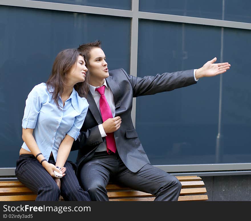 A young business man is showing something to his female colleague while sitting on a bench. Image taken outdoors, on an urban background. A young business man is showing something to his female colleague while sitting on a bench. Image taken outdoors, on an urban background.