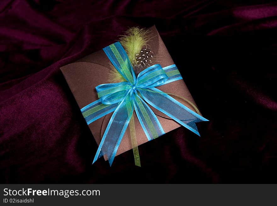 A brown gift folder with silk blue bow and guinea fowl feather on a maroon background. A brown gift folder with silk blue bow and guinea fowl feather on a maroon background