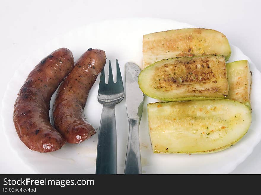 Two grilled sausages and zucchini on white plate with fork and knife between them. Two grilled sausages and zucchini on white plate with fork and knife between them