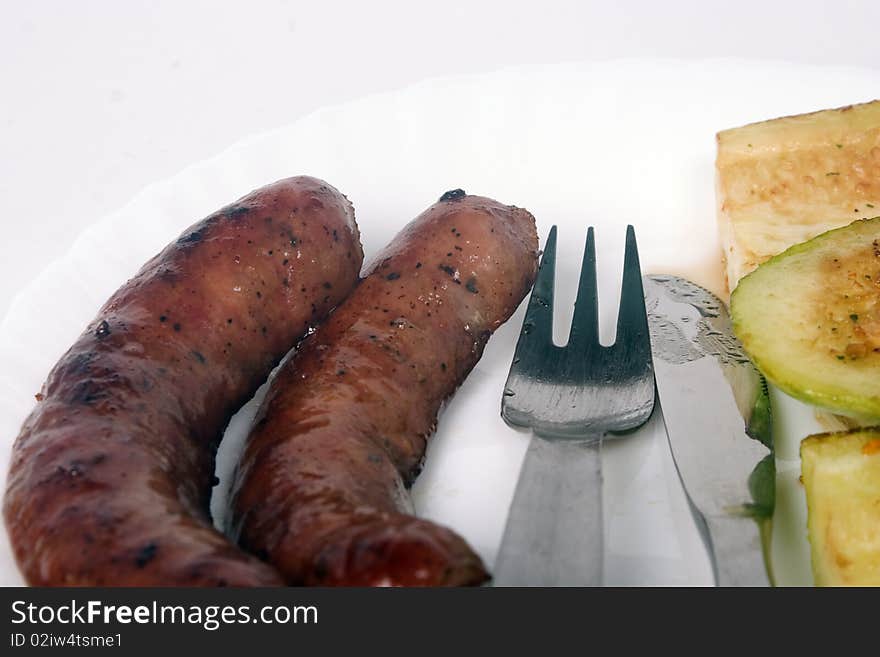 Two grilled sausages and zucchini on white plate with fork and knife between them. Two grilled sausages and zucchini on white plate with fork and knife between them