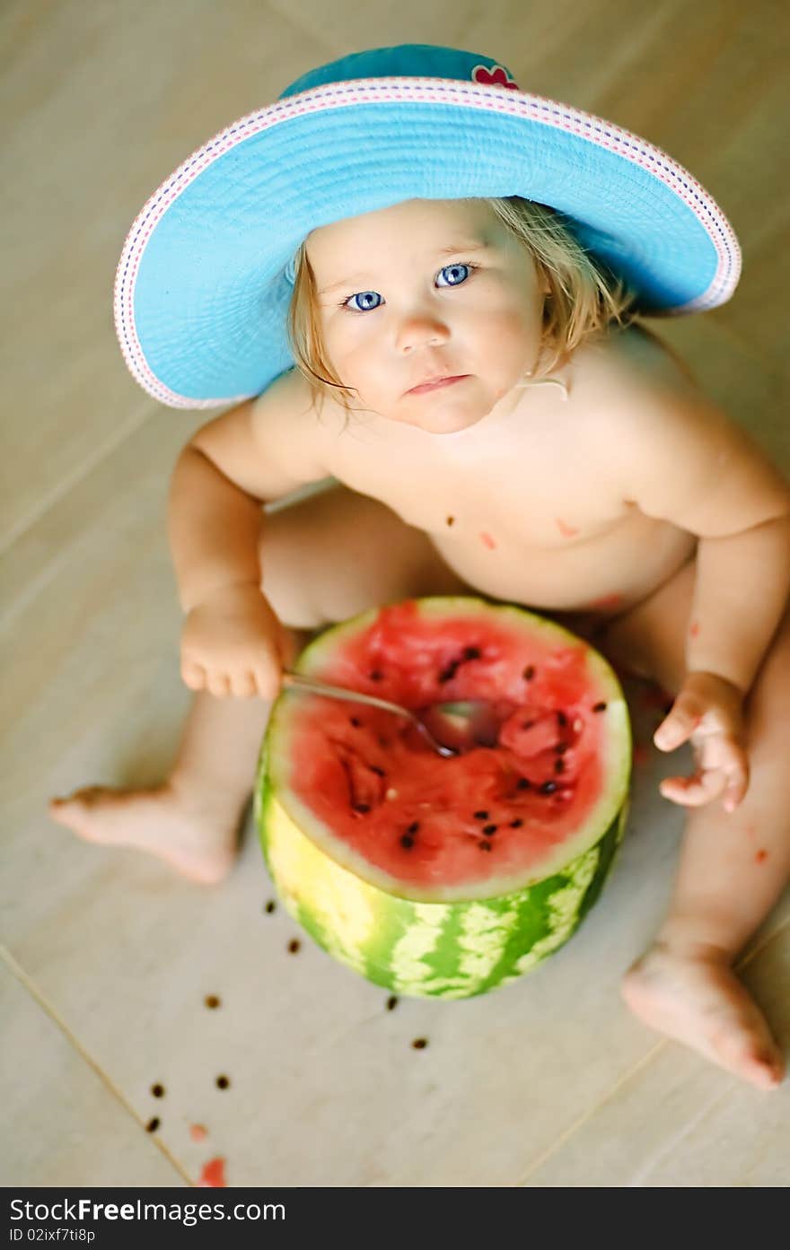 Cute baby-girl sits on a floor in a hat and eats a water-melon with a spoon. Cute baby-girl sits on a floor in a hat and eats a water-melon with a spoon