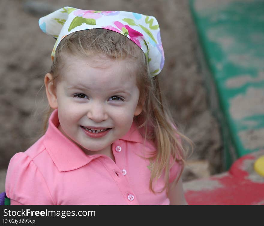 A Cute Girl Smiling in Sandbox with a Scarf on Head dressed in pink.