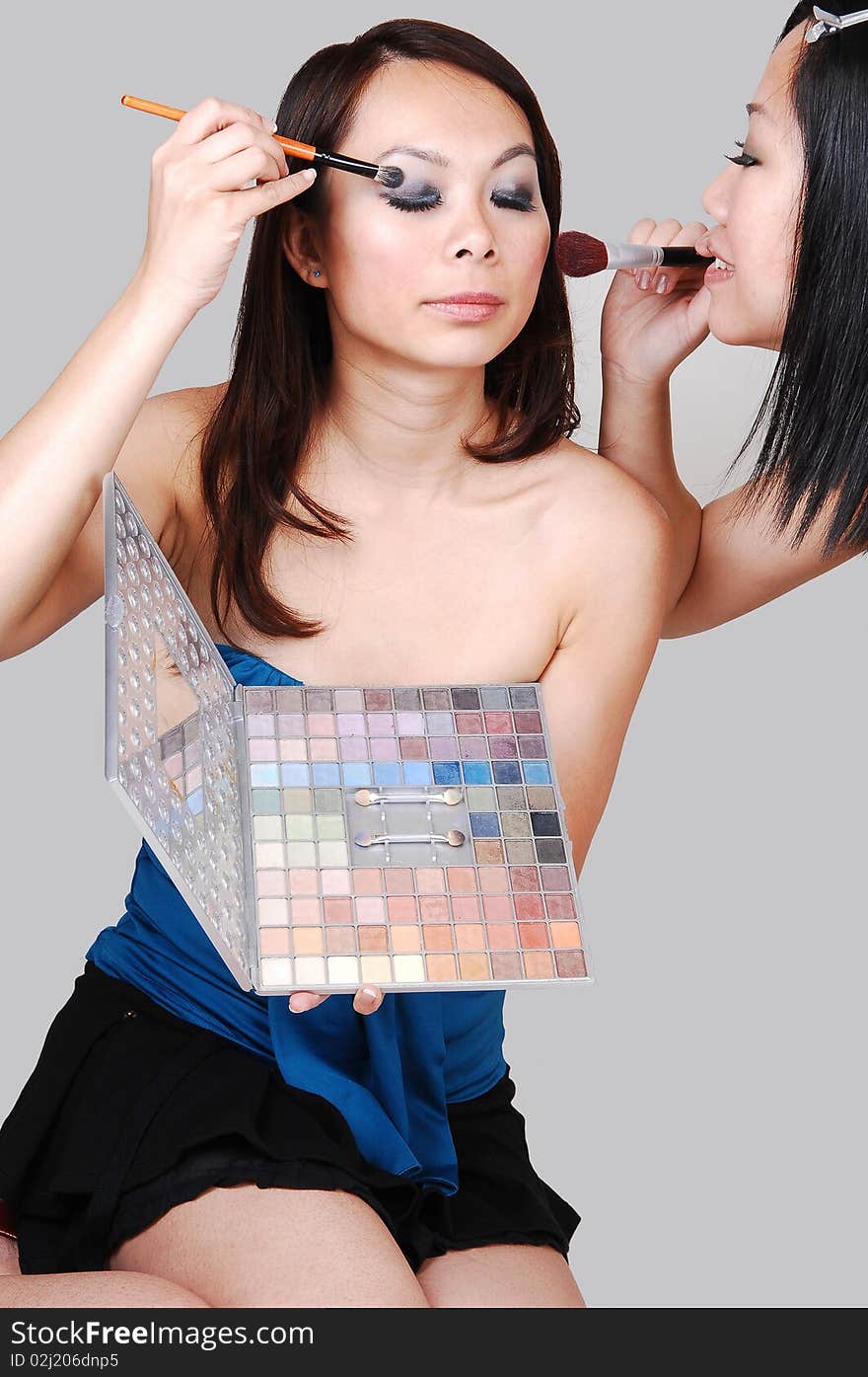 Two Asian woman kneeling on the floor in the studio and helping each other with the makeup for the next photo shoot, for light gray background. Two Asian woman kneeling on the floor in the studio and helping each other with the makeup for the next photo shoot, for light gray background.