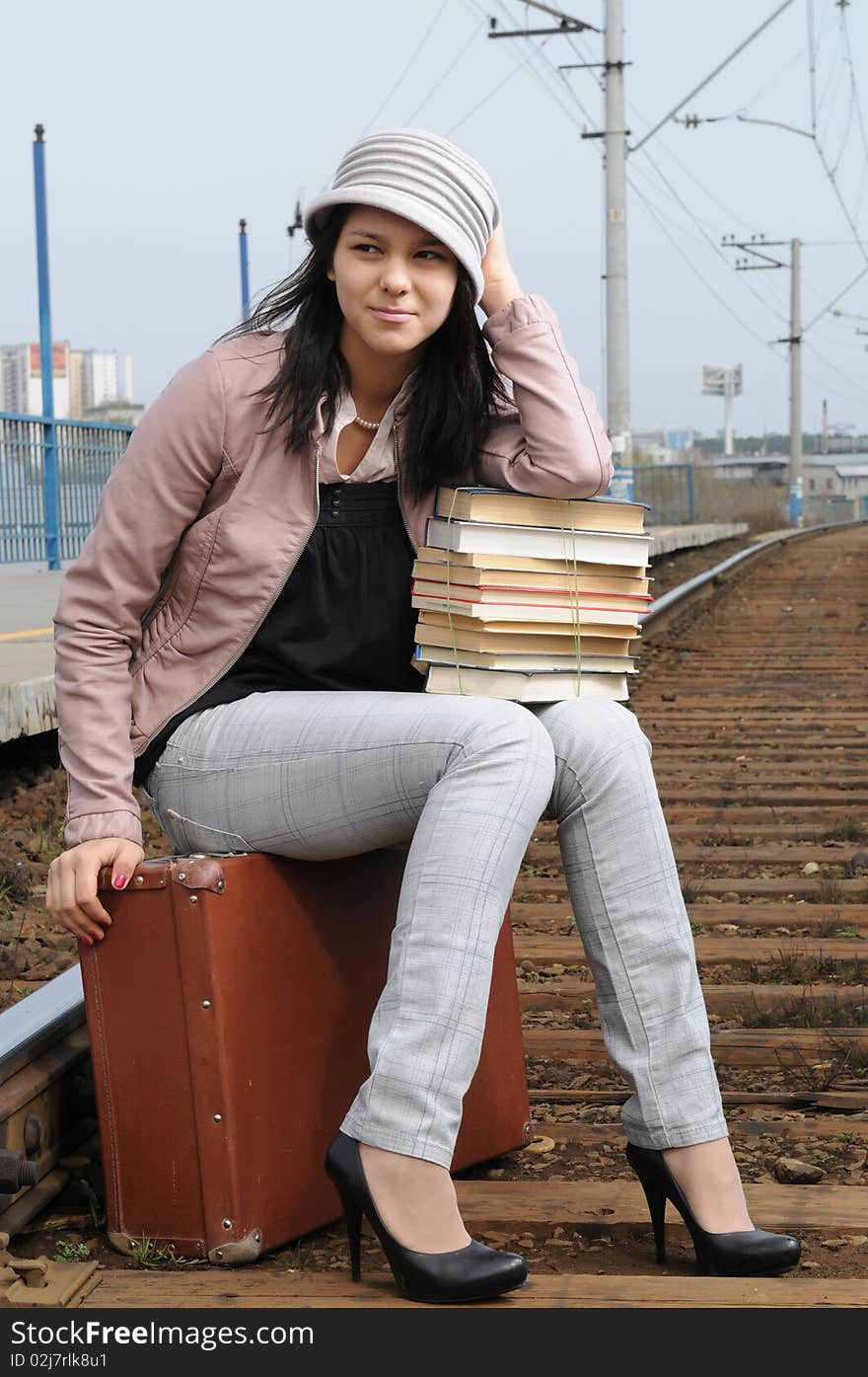 The young beautiful girl at station with a suitcase in a hand, in a light hat and a raincoat