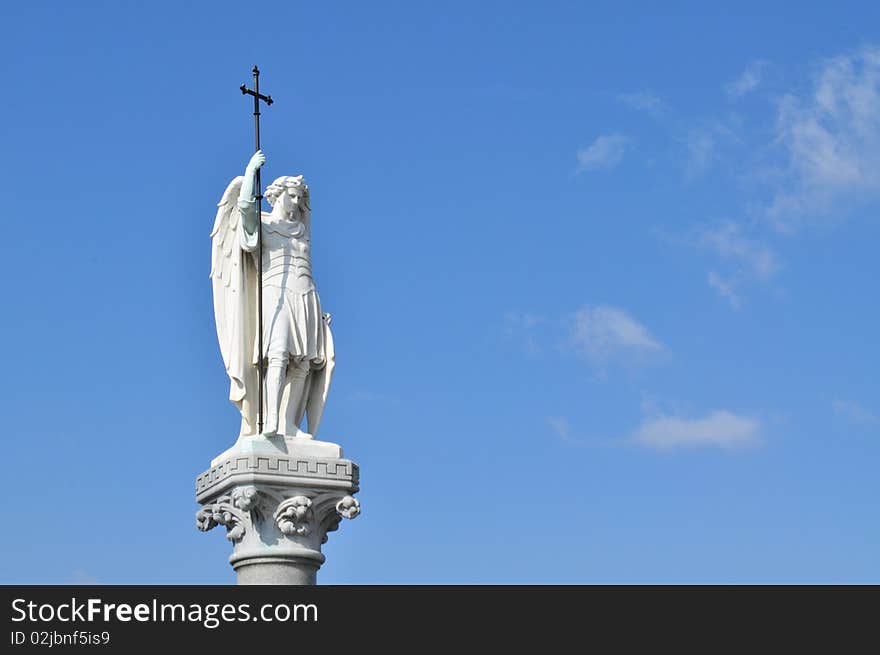 Statue of a winged holy man holding a cross
against a blue sky. Statue of a winged holy man holding a cross
against a blue sky