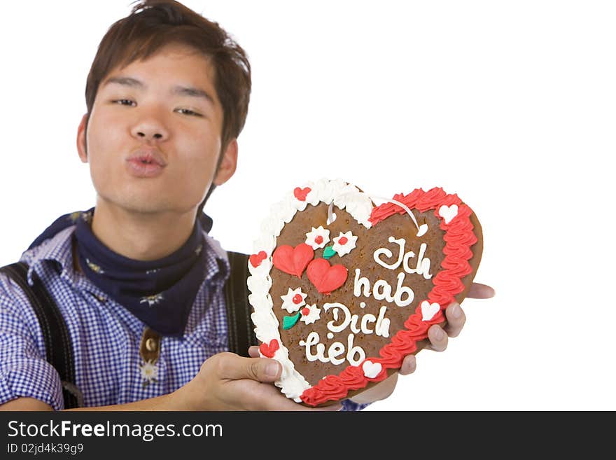 Chinese man, dressed with Bavarian Lederhose holds an Oktoberfest gingerbread heart in camera and gives a kiss. Isolated on white. Chinese man, dressed with Bavarian Lederhose holds an Oktoberfest gingerbread heart in camera and gives a kiss. Isolated on white.