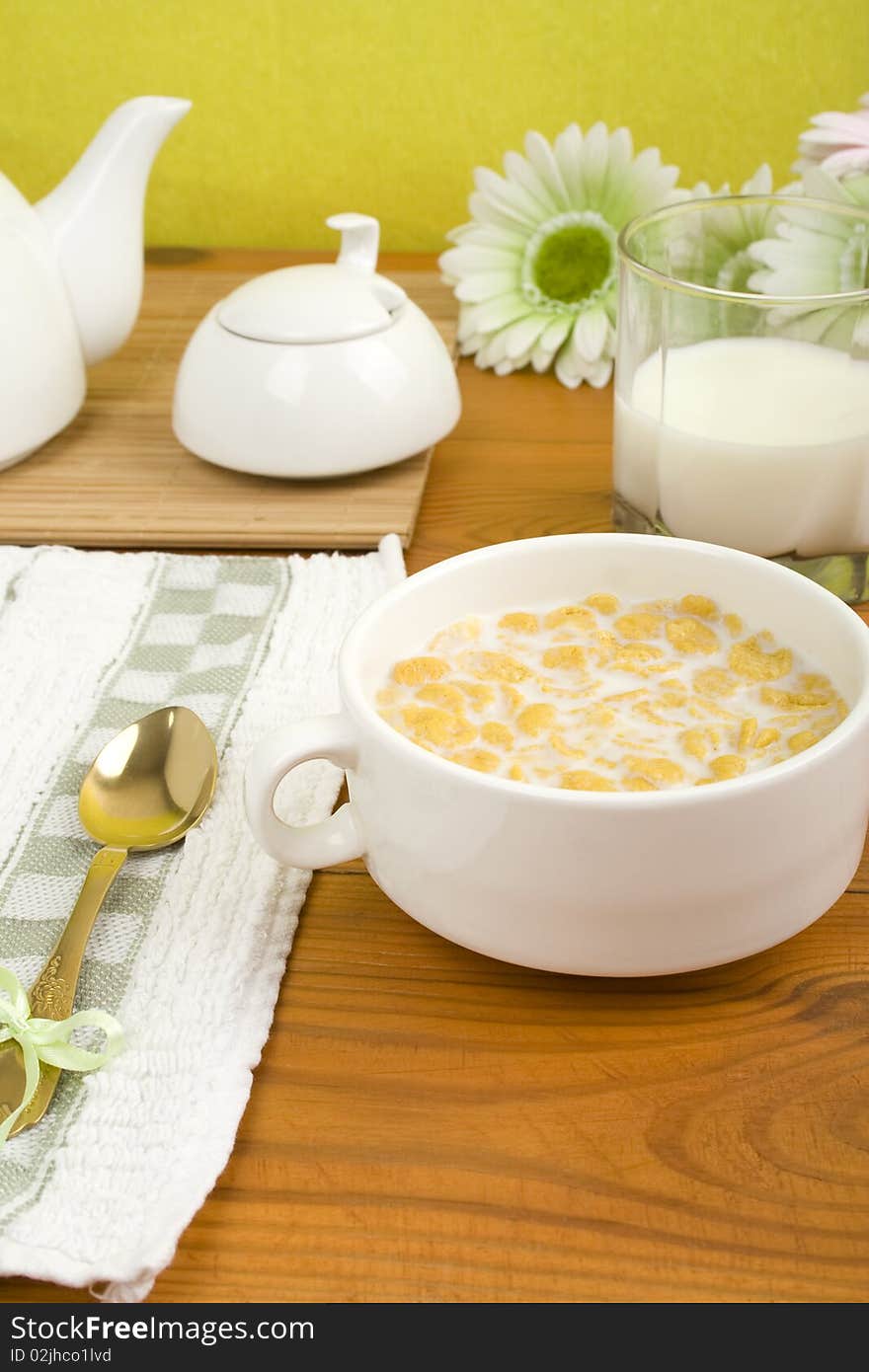 On the wooden table is breakfast cereal with milk, a glass of milk, a spoon, napkin, tea and flowers