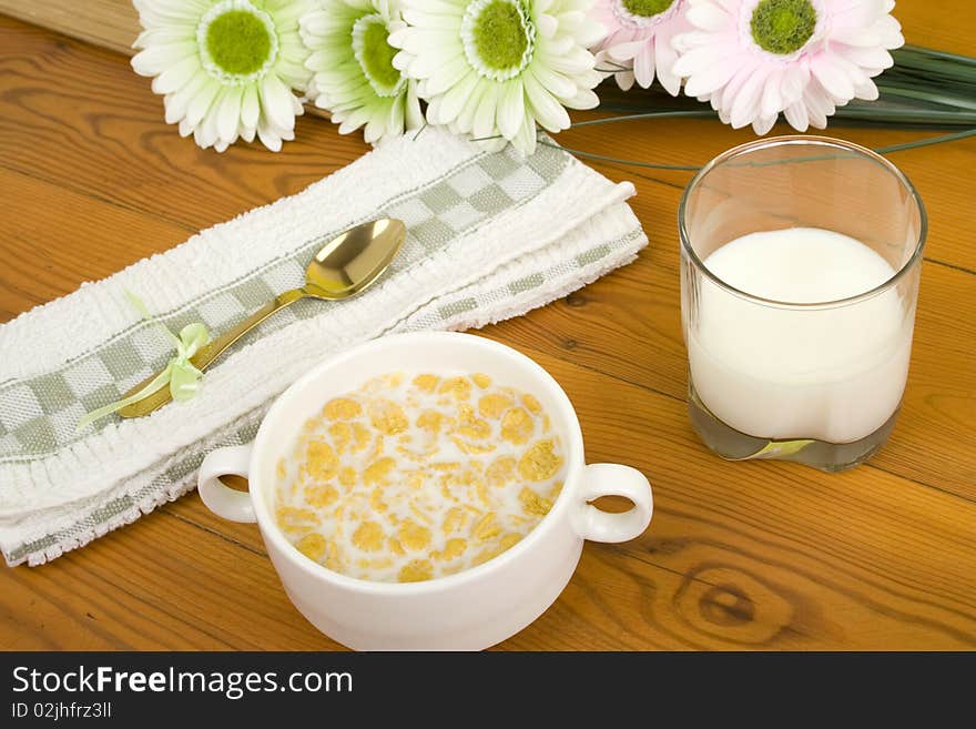 On the wooden table is breakfast cereal with milk, a glass of milk, a spoon, napkin, tea and flowers