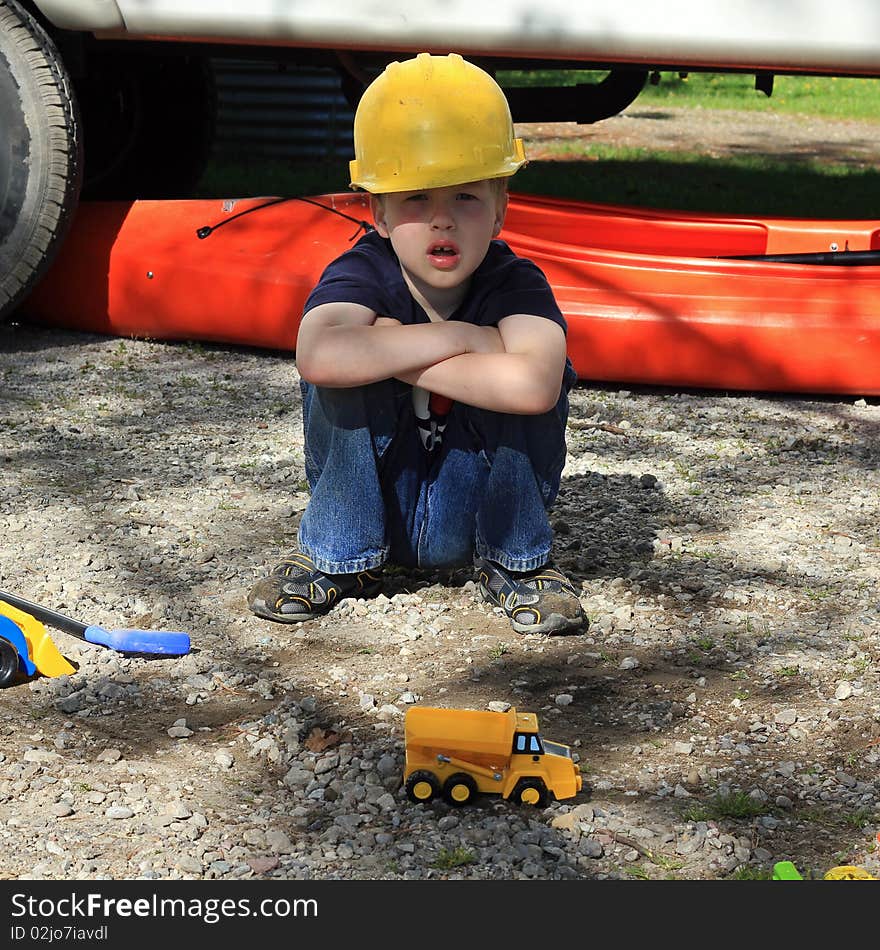 A young boy squatting on the ground wearing a yellow construction hard hat and playing with construction toys. He is at a campsite with an orange kayak in the background. A young boy squatting on the ground wearing a yellow construction hard hat and playing with construction toys. He is at a campsite with an orange kayak in the background.