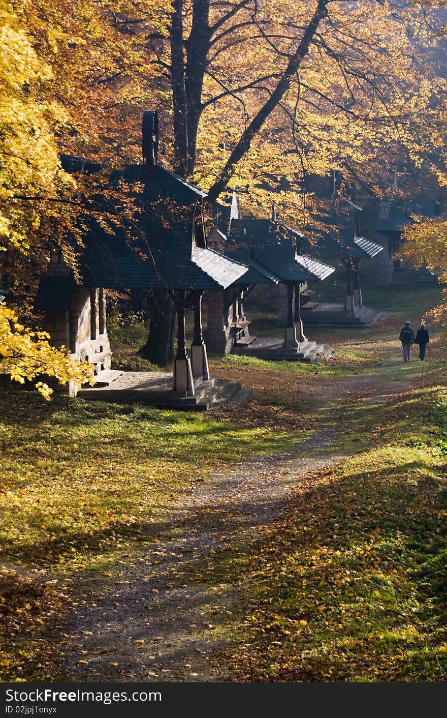 Sun shining through a trees along a walk way leading past small buildings (christian pilgrimage); a couple walking in the distance. Sun shining through a trees along a walk way leading past small buildings (christian pilgrimage); a couple walking in the distance.