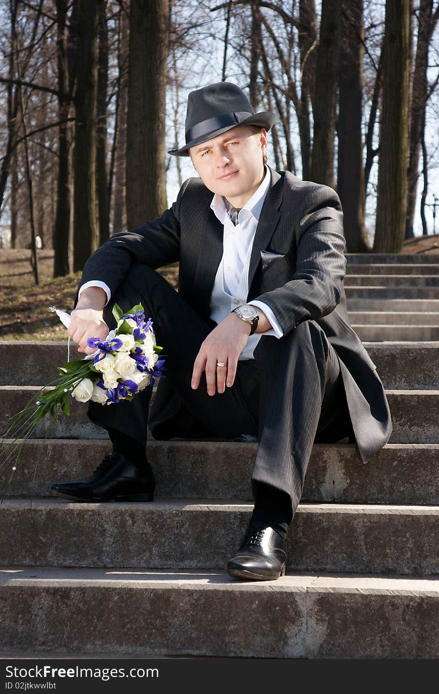 Young attractive smiling man, bridegroom, siting on stairs with bouquet, outdoor shot. Young attractive smiling man, bridegroom, siting on stairs with bouquet, outdoor shot