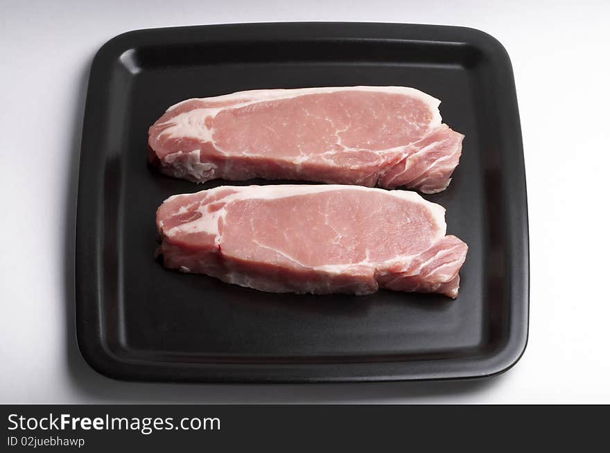 Two raw pork steaks on the black square plate over white background