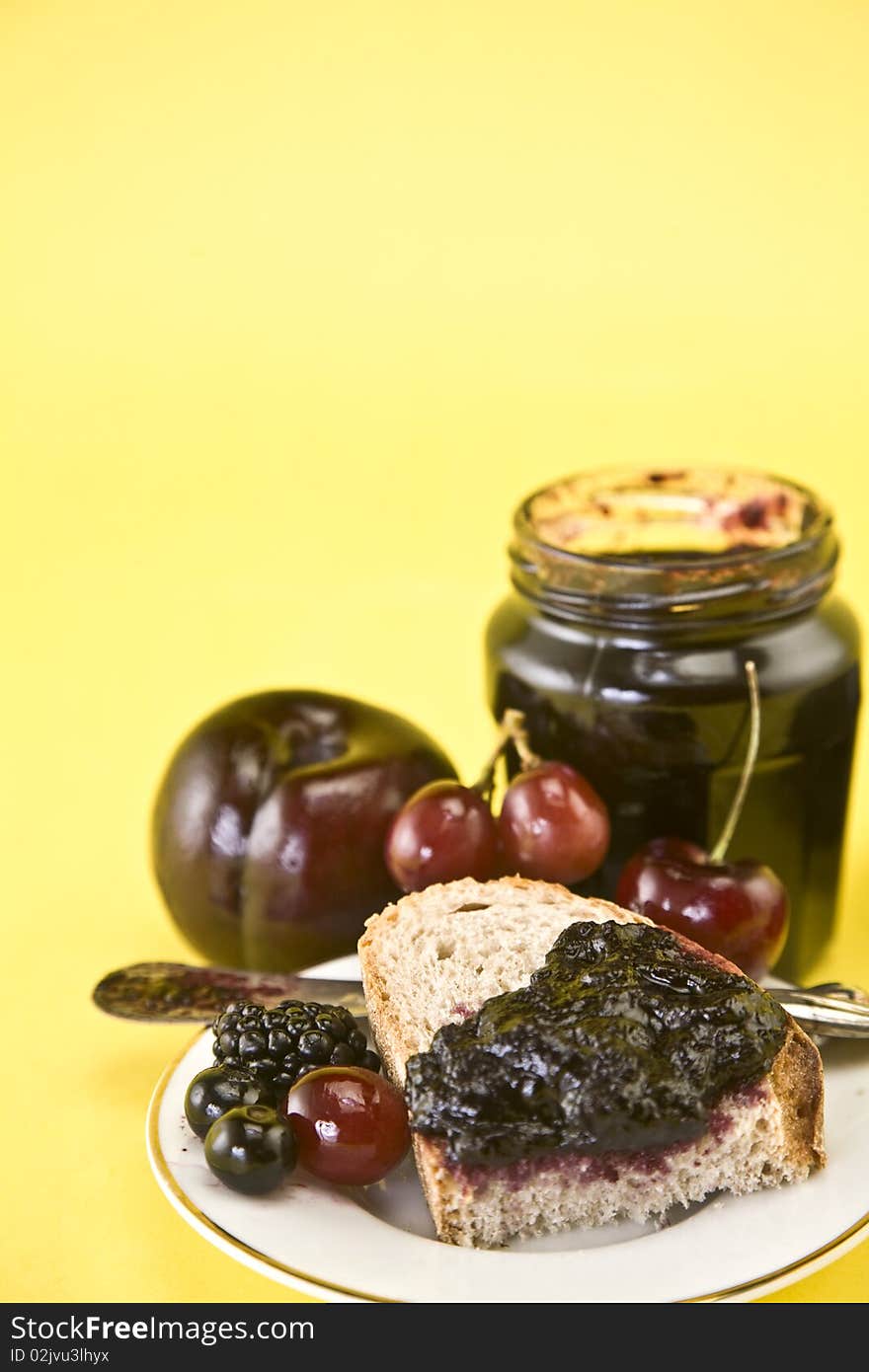 Blackberry jam in an open jar and on one piece of sliced bread on white plate against yellow background. Plum, cherries, blackberries and blueberries surround the jam. Blackberry jam in an open jar and on one piece of sliced bread on white plate against yellow background. Plum, cherries, blackberries and blueberries surround the jam.