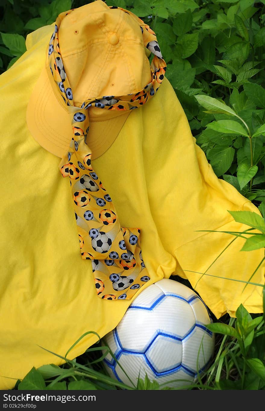 Yellow baseball cap, t-shirt, tie with soccer balls on it and soccer ball against green leaves. Yellow baseball cap, t-shirt, tie with soccer balls on it and soccer ball against green leaves.