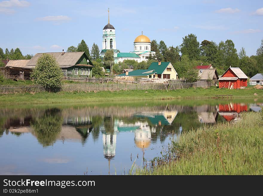 The image of an old orthodox church, rural wooden houses reflecting in the calm water of a river. The image of an old orthodox church, rural wooden houses reflecting in the calm water of a river