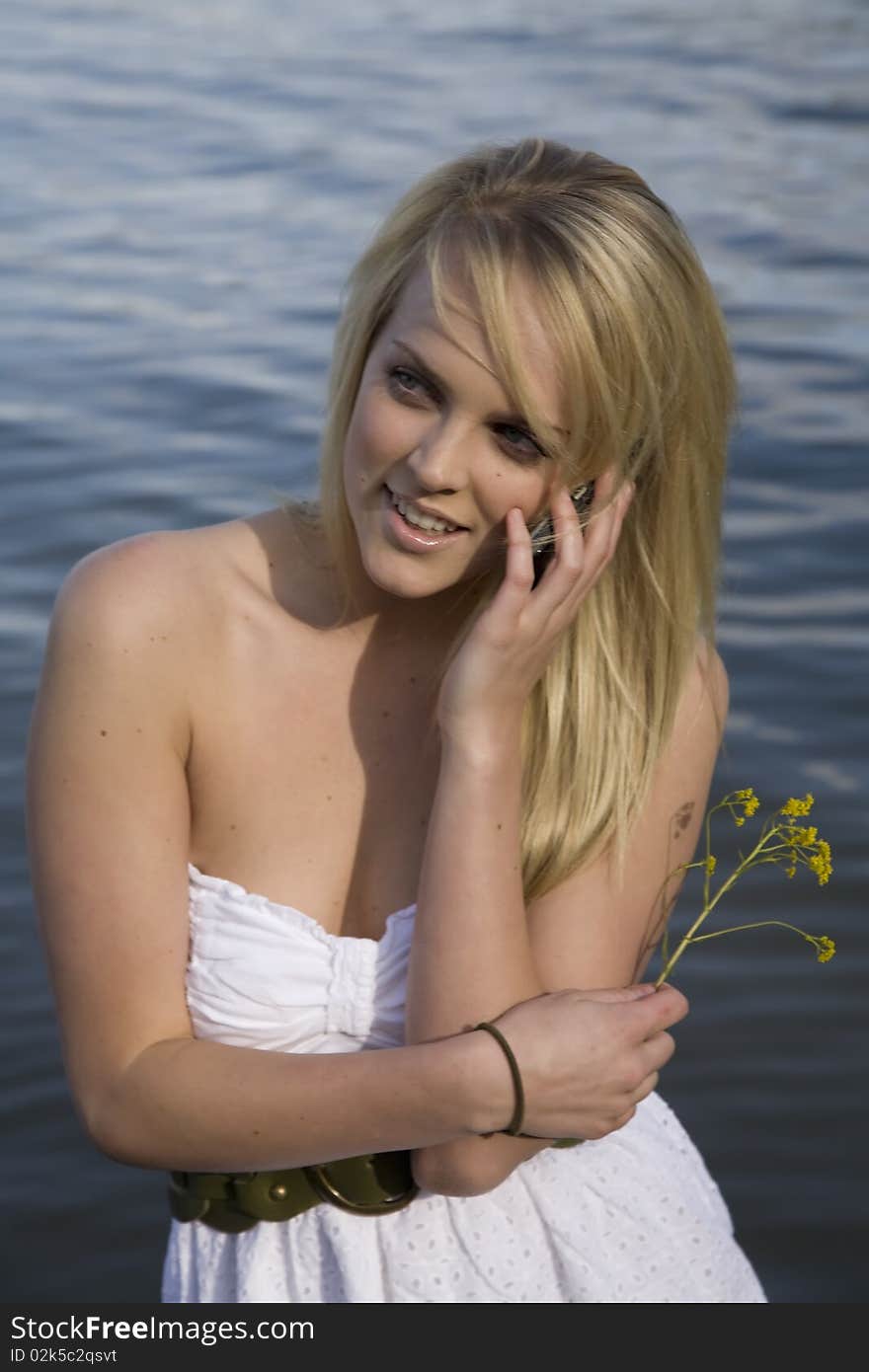 A woman in a white dress talking on the phone and standing in the water. A woman in a white dress talking on the phone and standing in the water.