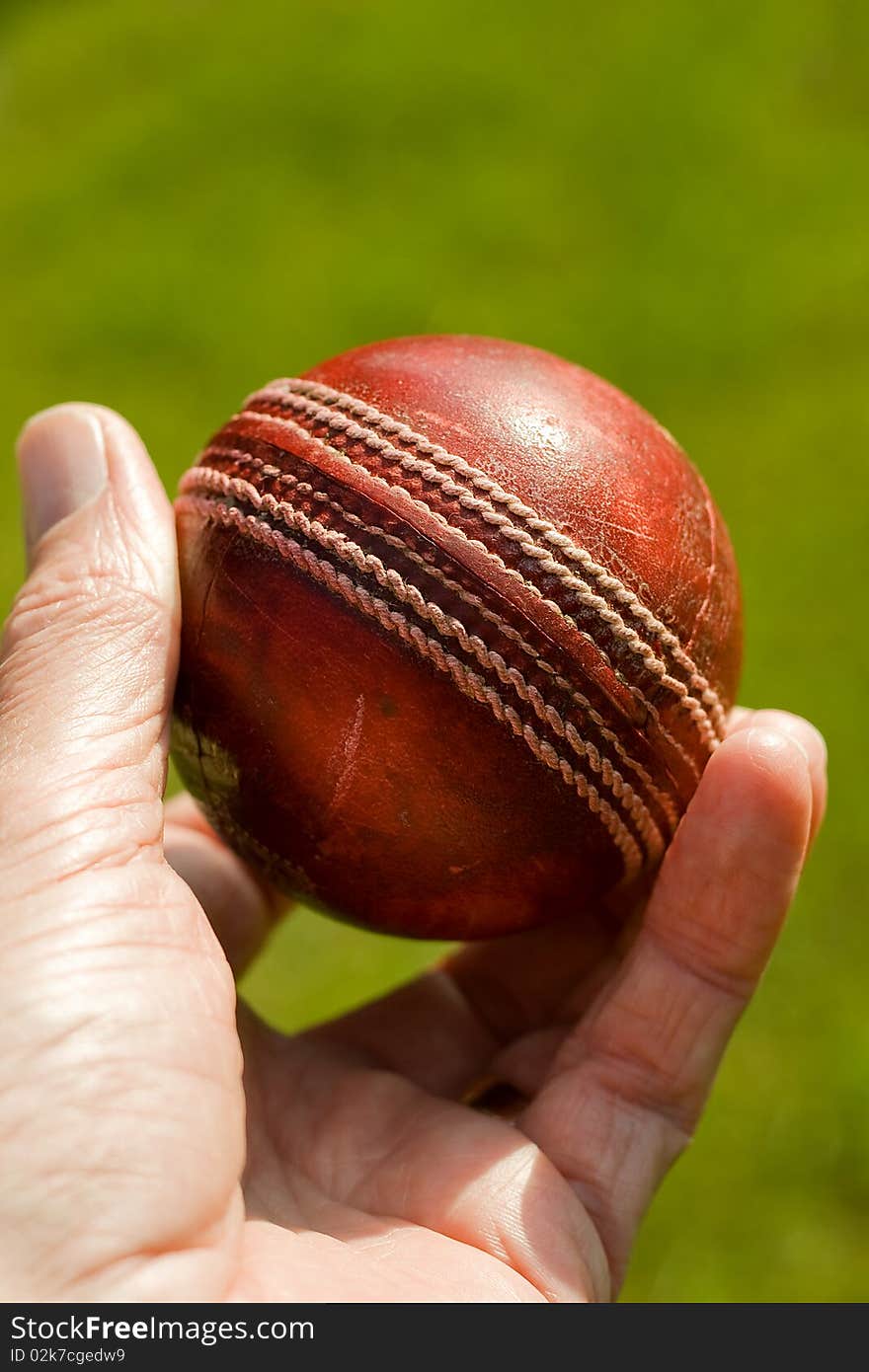 Used red leather cricket ball being held by hand against a green grass background