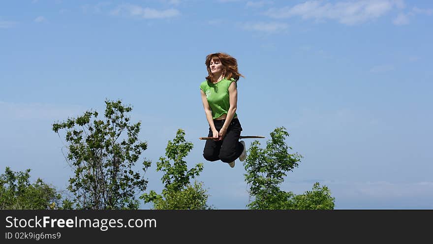 A young witch flies on a stick above trees.