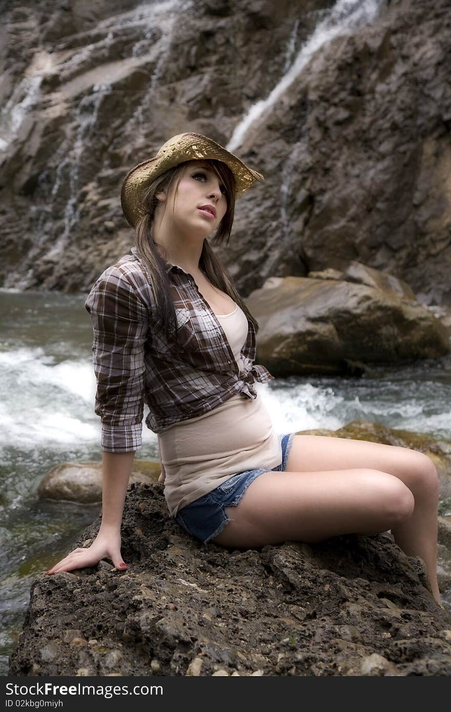 A young woman cowgirl sitting on a big rock by the river with waterfalls in the background. A young woman cowgirl sitting on a big rock by the river with waterfalls in the background.