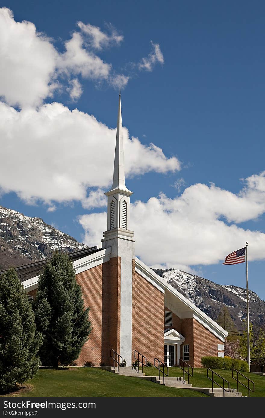 A small red brick church with a tall steeple and an American flag, with blue sky, snow-capped mountain and white clouds in the background. A small red brick church with a tall steeple and an American flag, with blue sky, snow-capped mountain and white clouds in the background