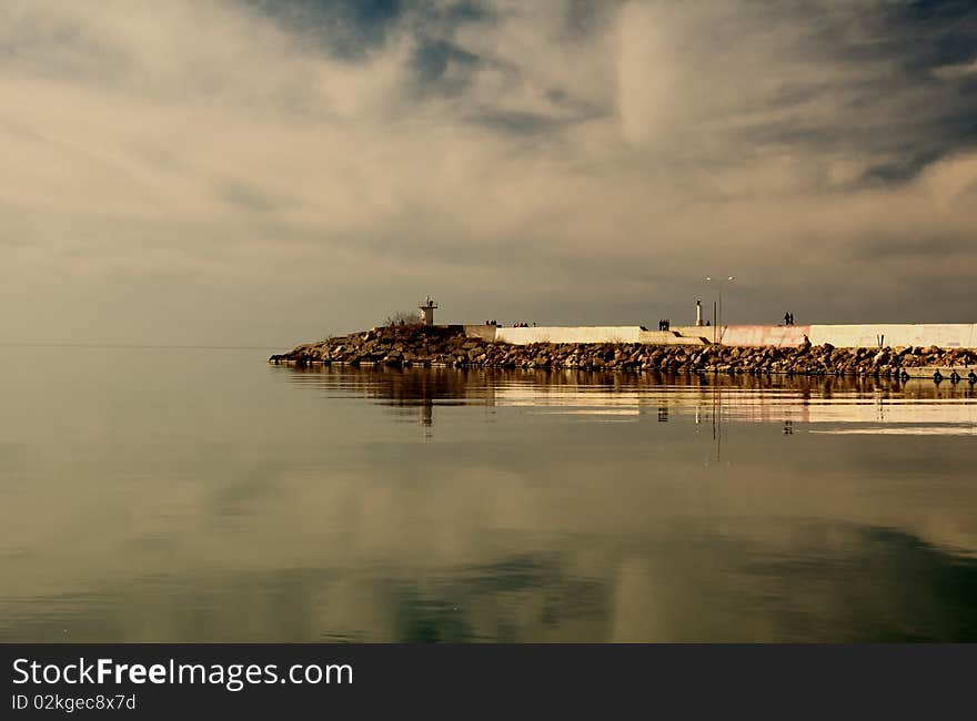 The mole (breakwater) with little lighthouse on black sea with clouds and the reflections of the clouds on the sea